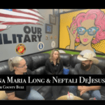 Three people are sitting and having a discussion on a black couch inside a room with military themed decorations and a pop art mural of a woman in sunglasses The text on the screen reads "Anna Maria Long & Neftali DeJesus Flagler County Buzz